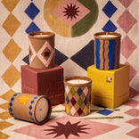 Colorful hand-painted ceramic candles with geometric patterns, displayed on vibrant boxes against a patterned backdrop.