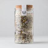 Glass jar with cork lid containing a mix of herbs or spices, on a light gray background.