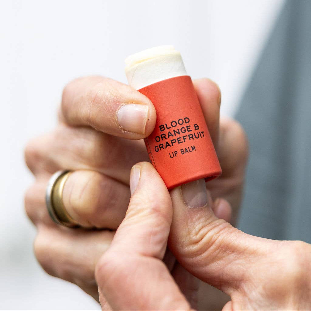 Red lip balm with 'Blood Orange & Grapefruit' flavor held in a hand against a light background