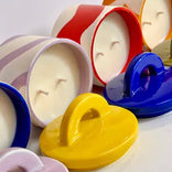 A group of colorful ceramic candle holders with matching lids, arranged closely together on a light background. The visible holders are white, red, blue, yellow, and lavender, each with a handle and a small candle inside.