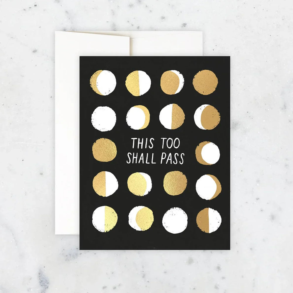 Greeting card with gold and white moon phases and text 'This Too Shall Pass' on a black background, placed on a marble surface.
