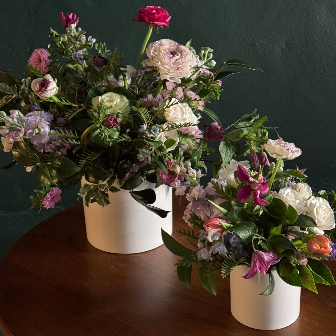Two flower arrangements in white vases on a wooden surface with a dark background