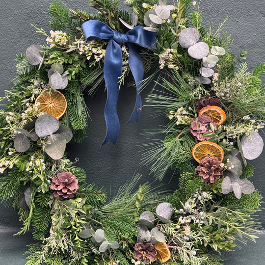 A picture of a holiday wreath with a blue ribbon, dried orange slices and pinecones against a dark green wall.