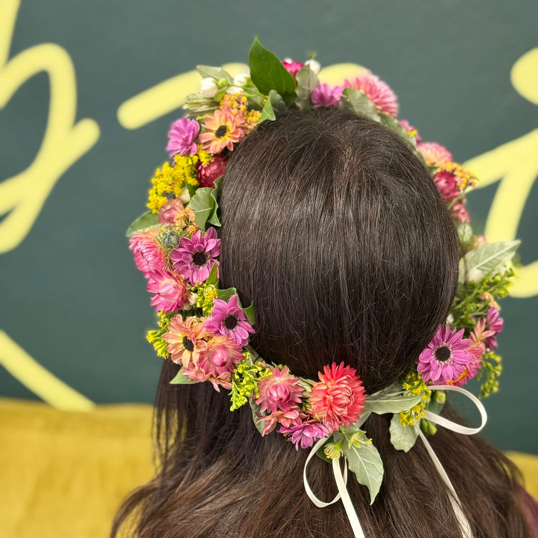 floral head crown sitting on a head of long brown hair. the crown is made up of purple, coral and yellow colored flowers.