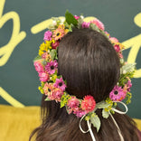 floral head crown sitting on a head of long brown hair. the crown is made up of purple, coral and yellow colored flowers.