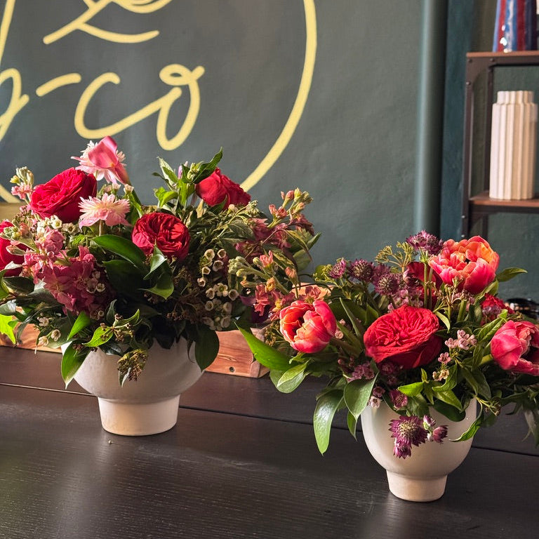 Two flower arrangements in white vases on a dark surface with a chalkboard background featuring a logo.