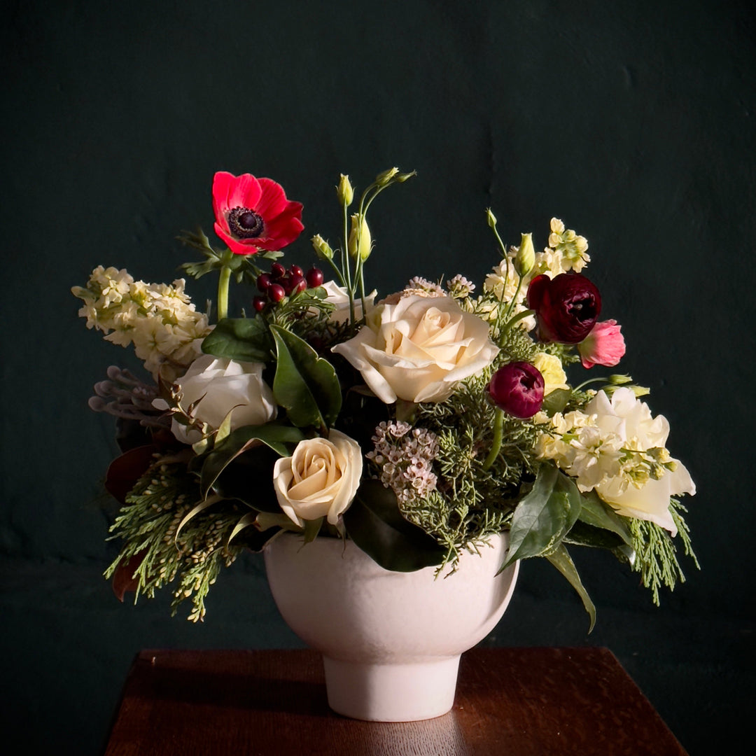 a white compote vase with a seasonal selection of flowers and greens arranged in a low table centerpiece style