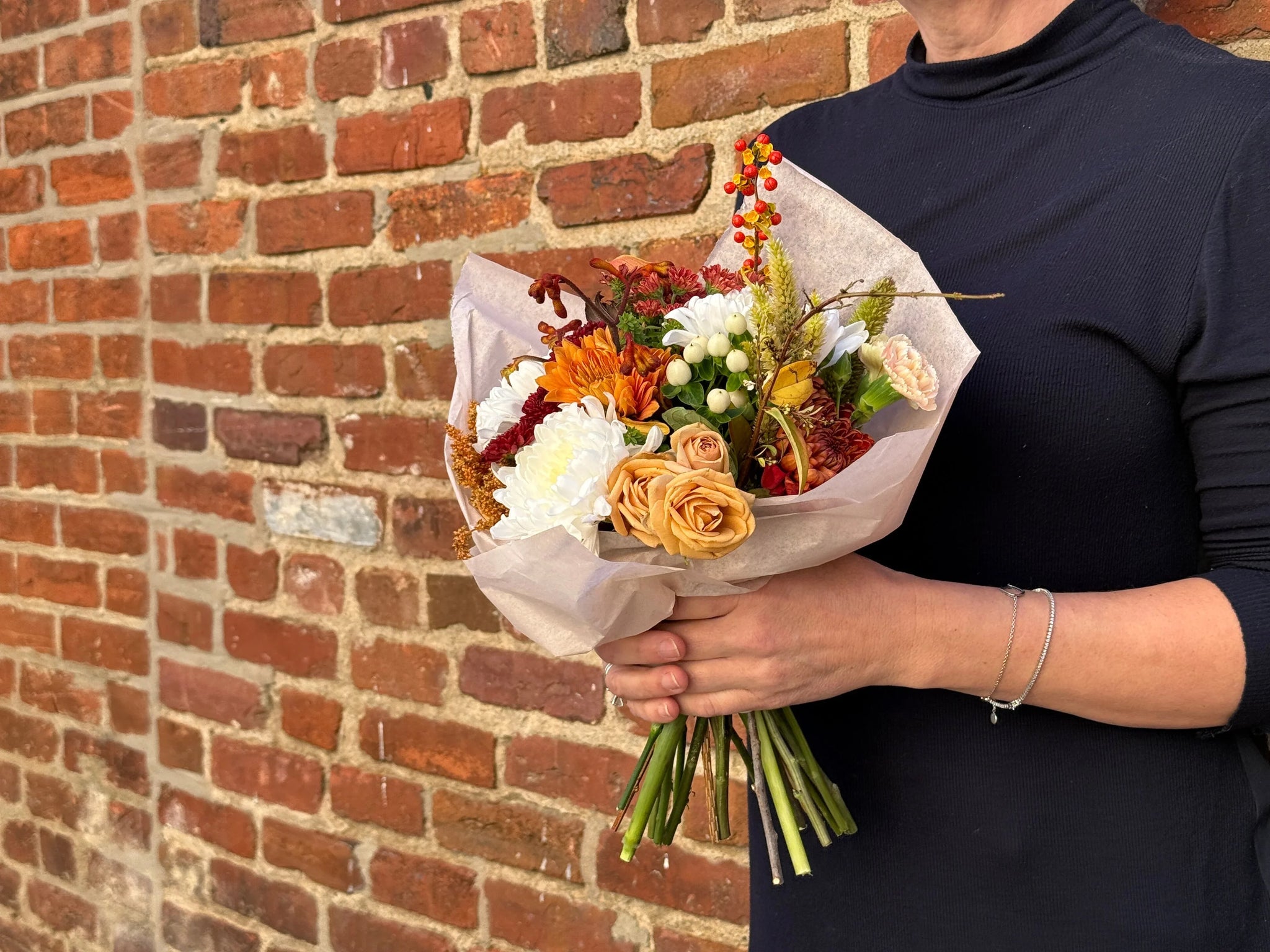 Autumn flower bouquet with orange roses, white blooms, and berries, held against a brick wall.