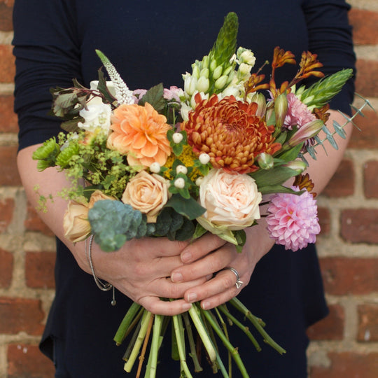 large flower bouquet with a mix of seasonal flowers in soft pinks and apricots and foliage being held by two hands. 