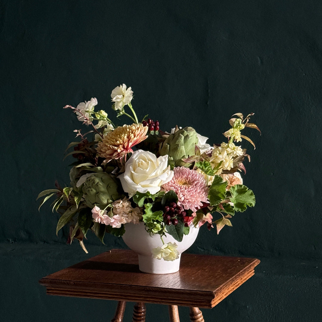 A floral arrangement with a mix of fall inspired blooms in a soft color palette, displayed in a white vase, placed on a wooden table against a dark background.