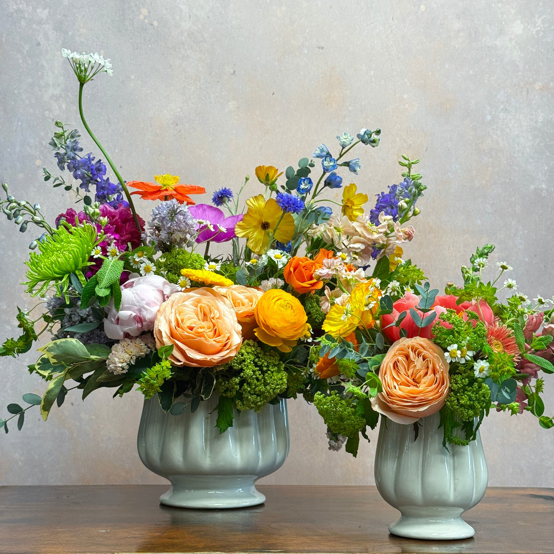 vases, one large and one small of colorful flowers sitting on a wooden table.