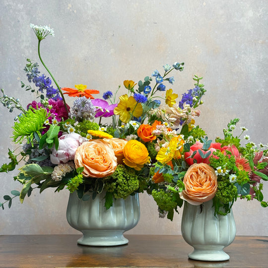 vases, one large and one small of colorful flowers sitting on a wooden table.