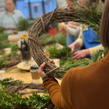 Person crafting a wreath with greenery at a table with other materials.