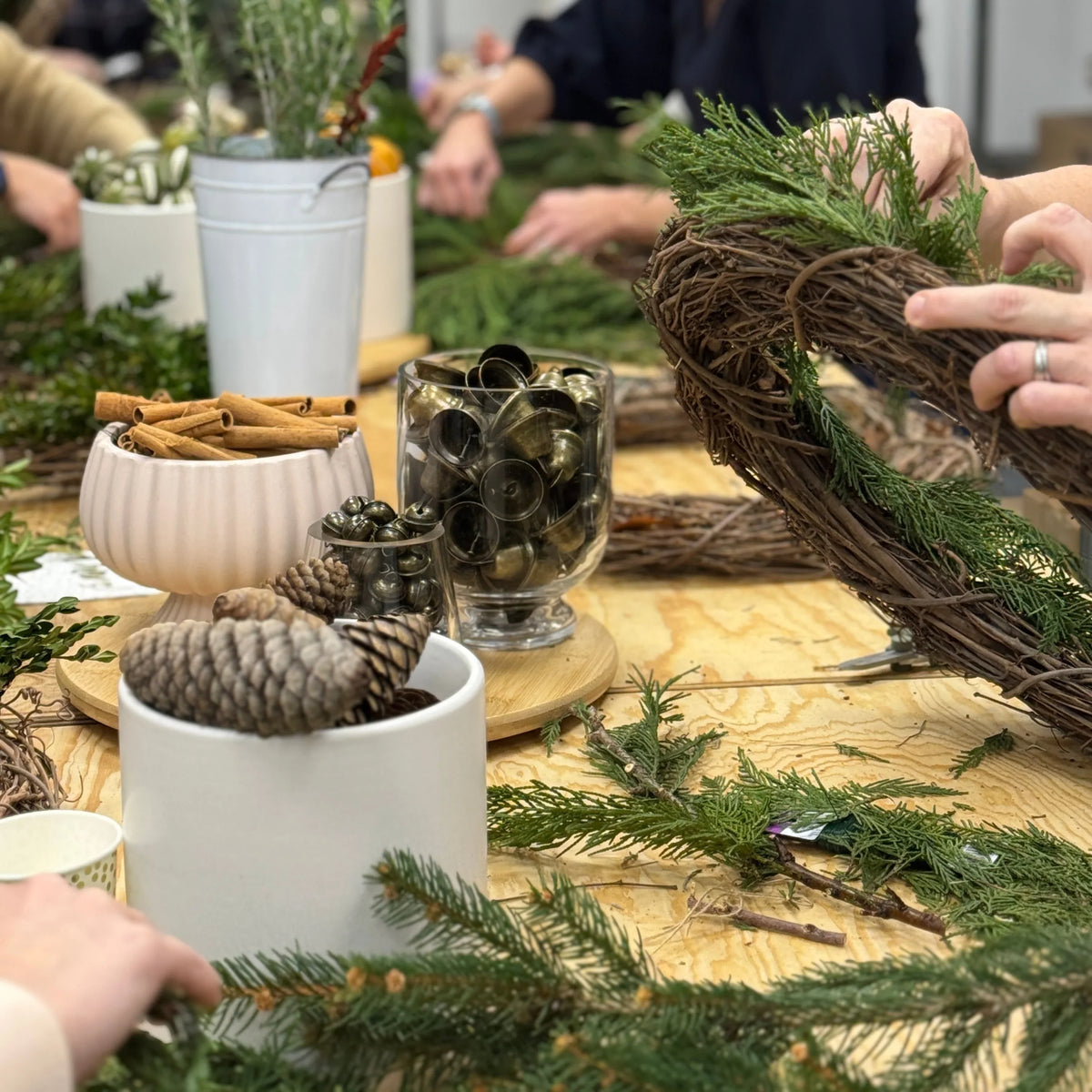 Hands crafting a festive wreath with pine, cinnamon, and jingle bells at a floral workshop.