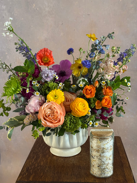 brightly colored flower arrangement in a white vessel sitting on a table next to a jar of bath salts