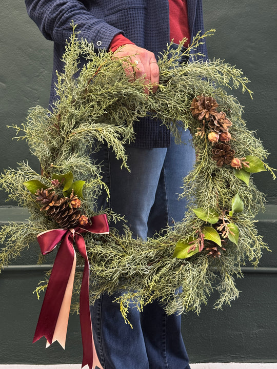 Person holding a festive wreath with greenery, pinecones, and a red ribbon against a dark background.