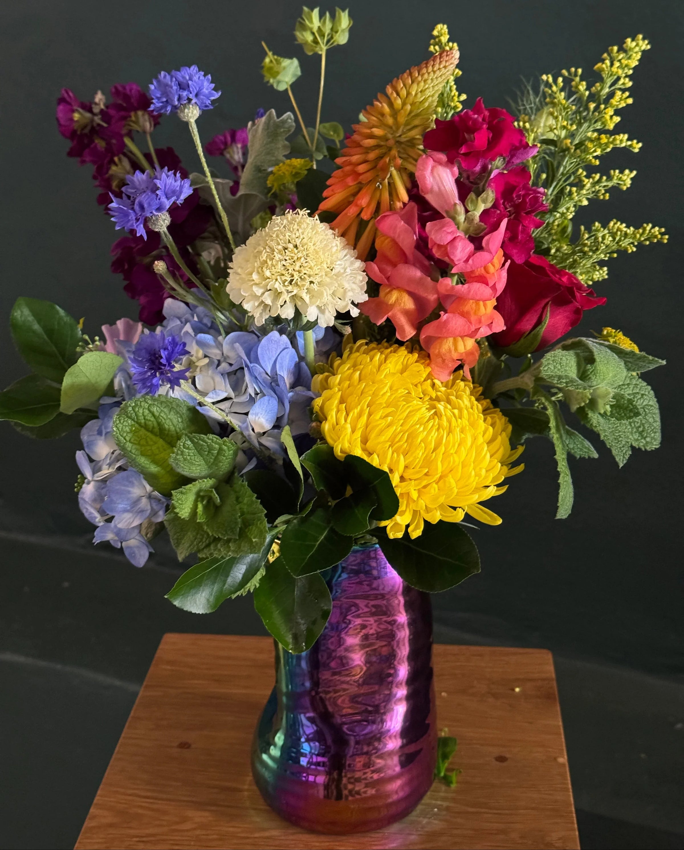 Colorful bouquet of flowers in a textured vase on a wooden surface with a black background