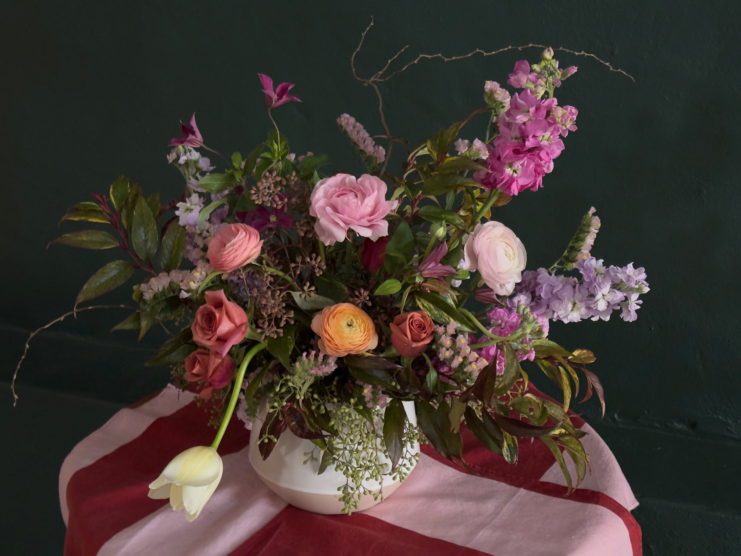 Floral arrangement in a white ceramic vase set on a pink and red striped table cloth. Flowers are soft pinks and purples. 