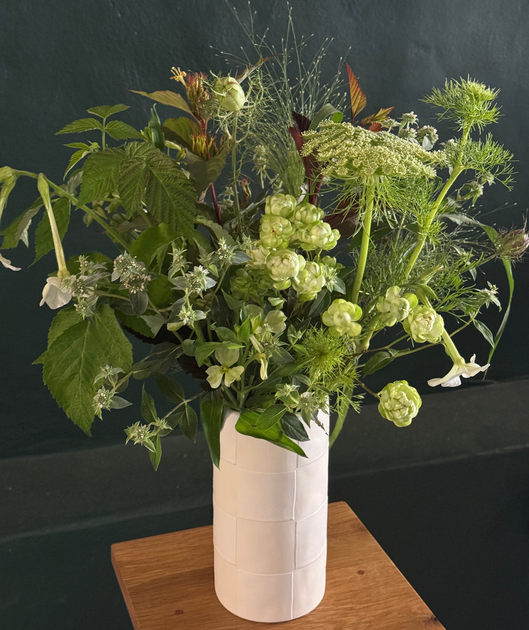A lush green and white floral arrangement in a textured white ceramic vase, placed on a wooden block against a dark green wall.