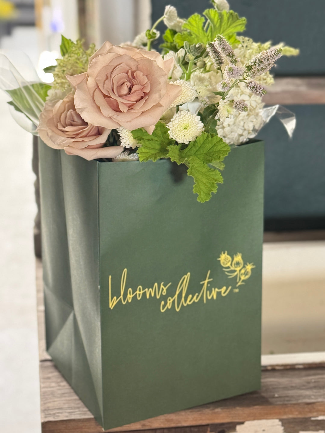 A green paper gift bag with gold foil script reading 'blooms collective' filled with a floral arrangement of blush pink roses, white flowers, and green foliage, set on a rustic wooden surface.