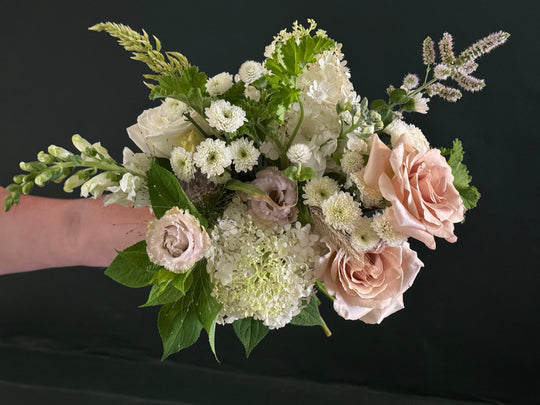 A floral bouquet held by a person's hand against a dark black background, featuring blush pink roses, white hydrangeas, daisies, and green foliage.