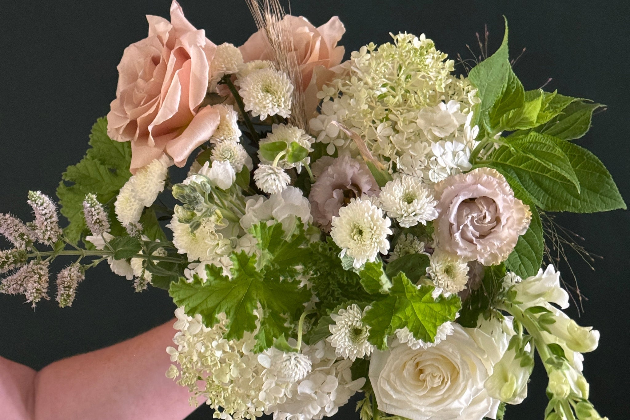 A hand holding a lush bridal bouquet featuring blush pink and white roses, white chrysanthemums, green foliage, and delicate wildflowers against a dark background.