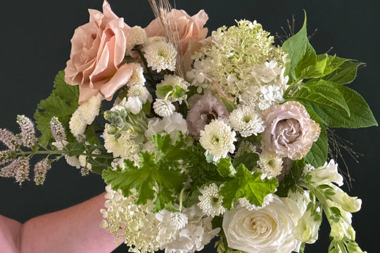 A hand holding a lush bridal bouquet featuring blush pink and white roses, white chrysanthemums, green foliage, and delicate wildflowers against a dark background.