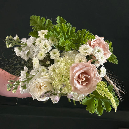 A floral bouquet featuring blush pink roses, white chrysanthemums, and green foliage, held by a person whose arm is partially visible against a dark background.