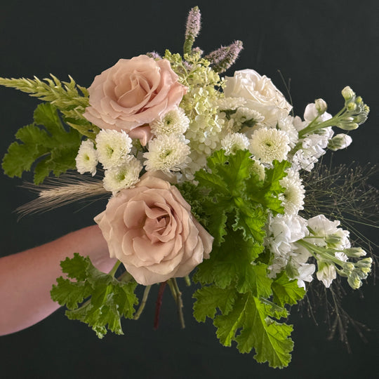 A hand holding a rustic-style floral bouquet featuring blush pink roses, white chrysanthemums, green foliage, and wild grasses against a dark background.