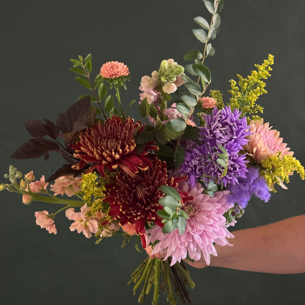 A hand holding a vibrant, multicolor flower bouquet featuring pink, purple, burgundy, and yellow flowers with green foliage, set against a dark green background.
