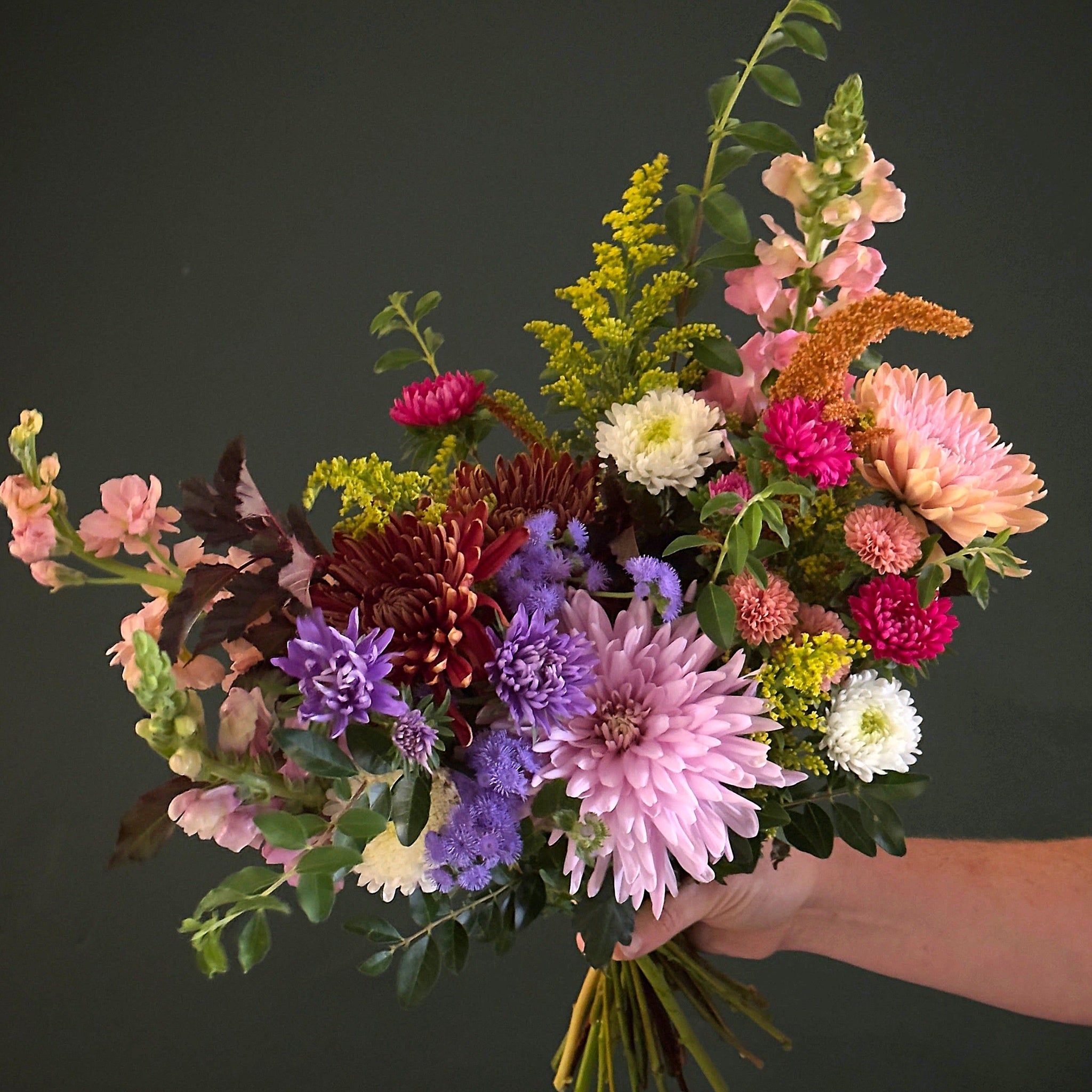 A vibrant mixed flower bouquet held by a hand against a dark gray background. The bouquet features a variety of flowers including light pink chrysanthemums, purple and white asters, solidago and pink snapdragons with assorted greenery.