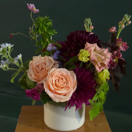 A floral arrangement featuring peach roses, deep burgundy chrysanthemums, and green foliage in a white ceramic pot, set on a wooden surface against a dark green background.