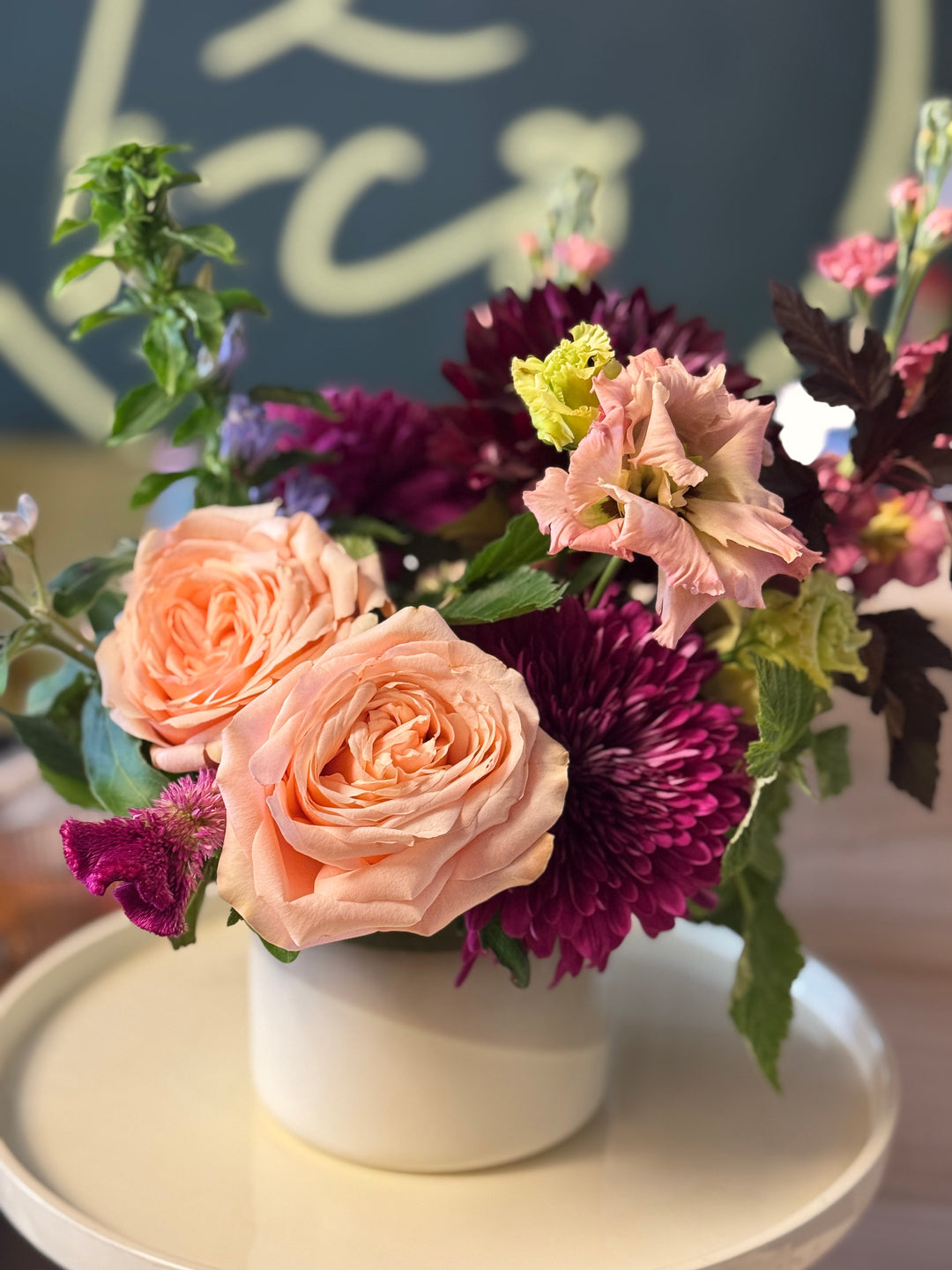 A floral arrangement of peach roses, deep magenta chrysanthemums, and light pink flowers with green foliage, presented in a small white cylindrical vase on a round white tray.