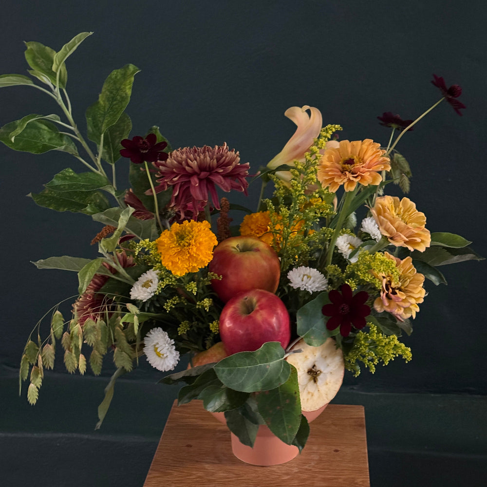 An autumnal floral arrangement in a terracotta pot, featuring orange and yellow flowers, burgundy dahlias, white daisies, green foliage, and red apples, displayed on a wooden pedestal against a dark teal background.
