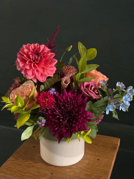 A vibrant mixed flower arrangement in a white ceramic pot, featuring a large pink dahlia, a deep purple chrysanthemum, peach roses, and blue flowers, set on a wooden surface against a dark background.