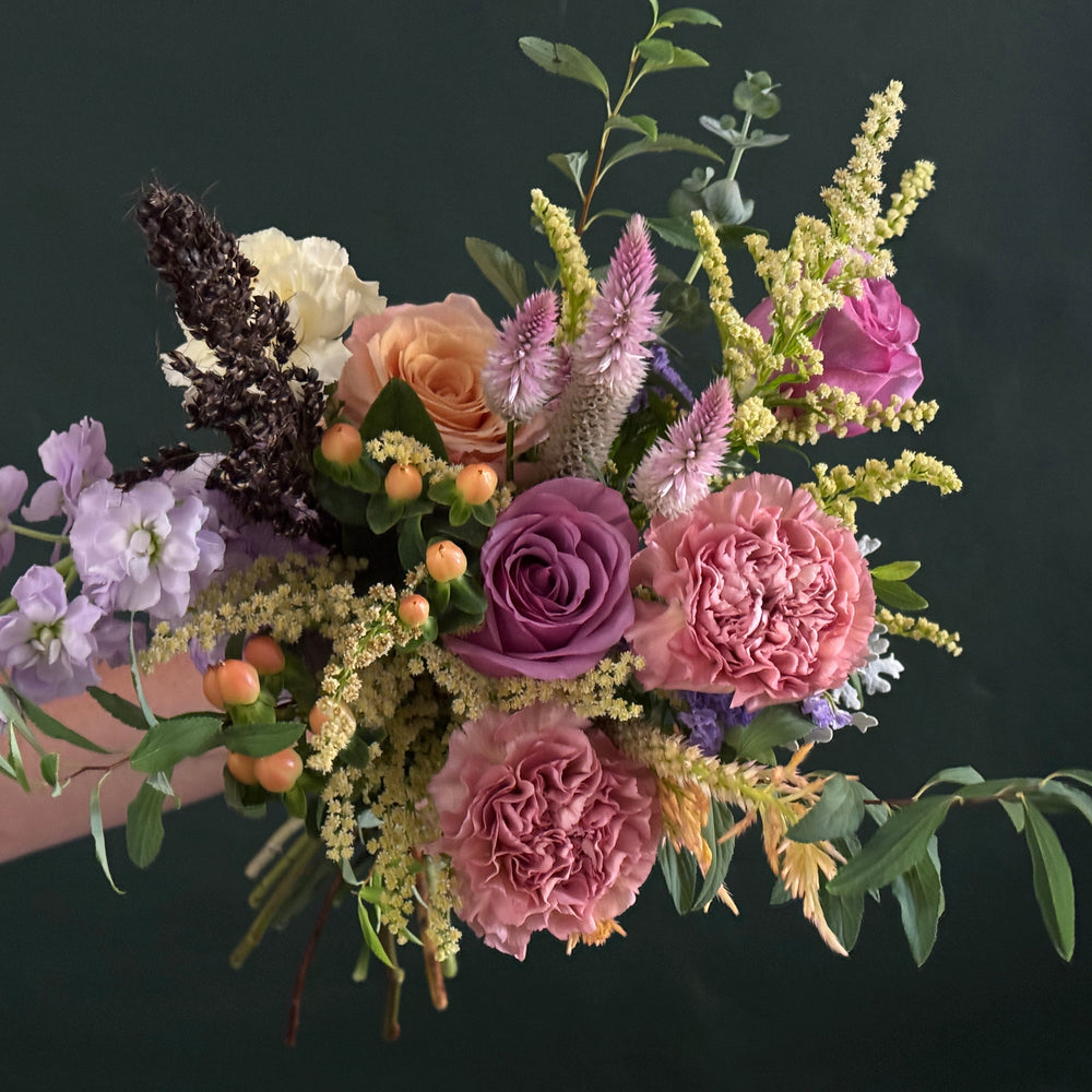 A hand holding a lush, rustic-style floral bouquet featuring pink and purple roses, lavender, peach roses, thistle, and green foliage against a dark green chalkboard background.