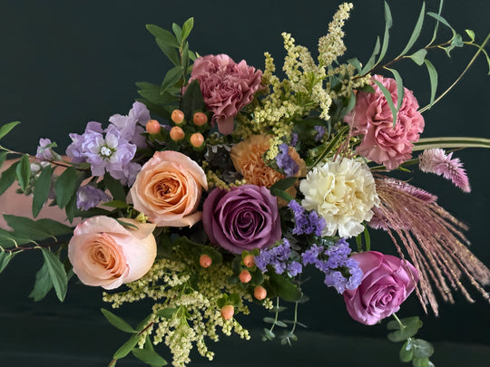 A lush floral arrangement featuring peach roses, purple and pink carnations, lavender sprigs, cream flowers, green foliage, and dried grasses, set against a dark green background.