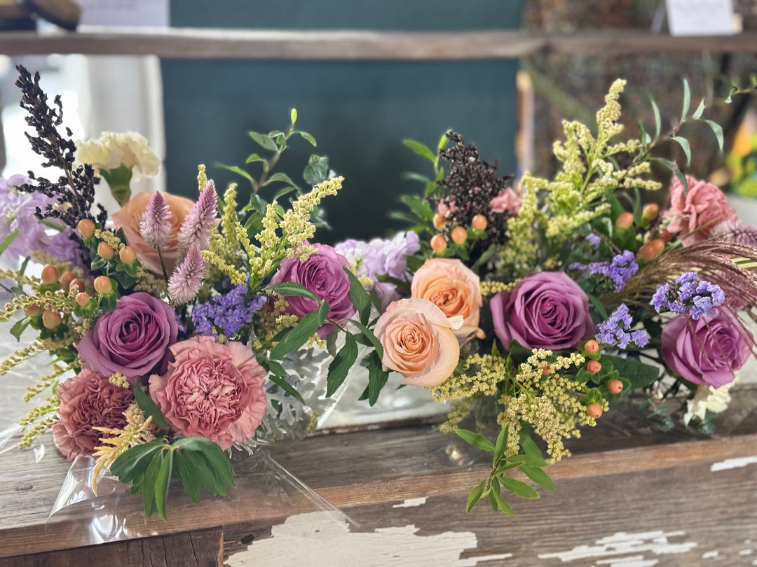 Two rustic floral arrangements in clear glass vases on a weathered wooden table, featuring purple roses, peach roses, pink peonies, and green foliage.