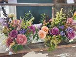 Two rustic floral arrangements in clear glass vases on a weathered wooden table, featuring purple roses, peach roses, pink peonies, and green foliage.