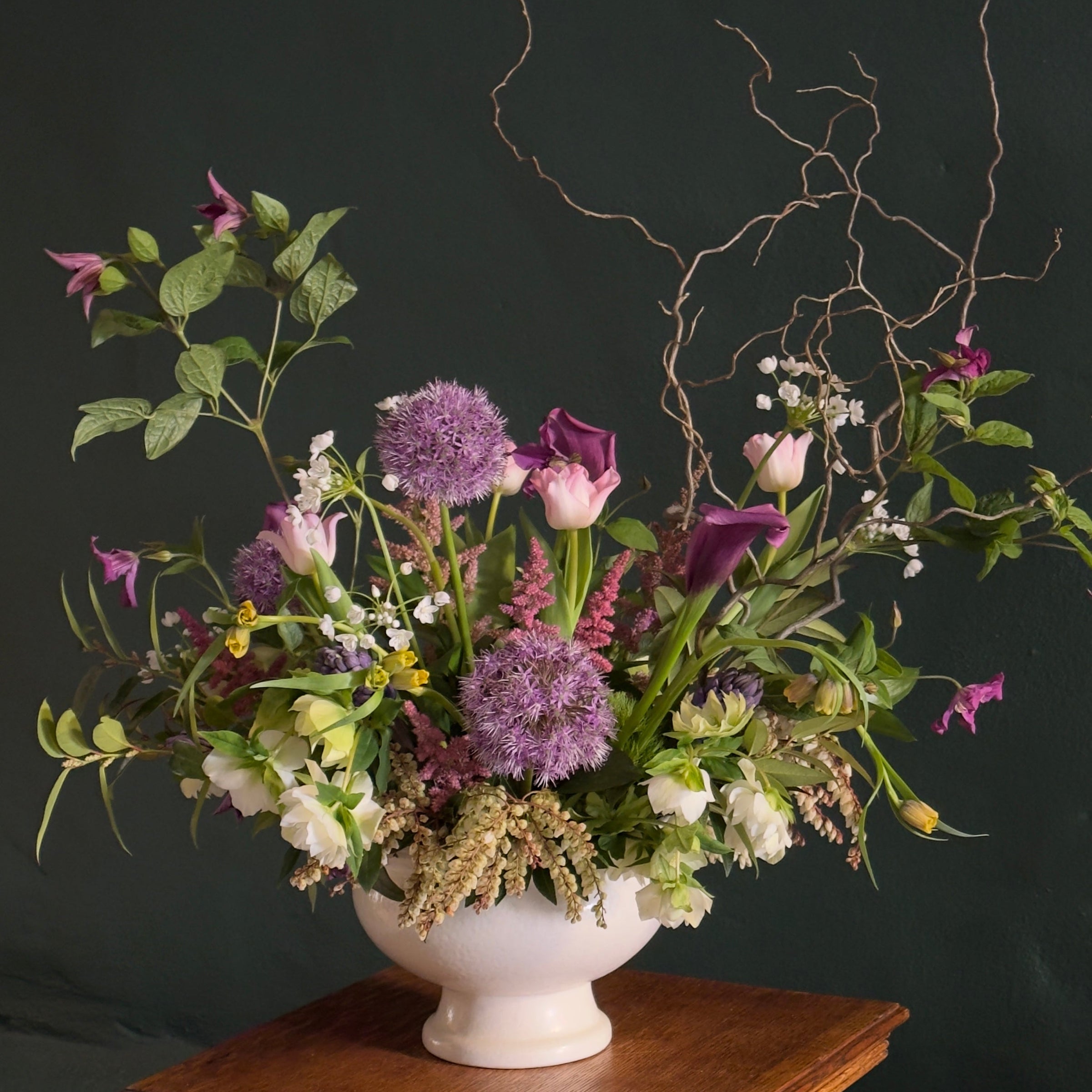 A floral arrangement featuring a variety of flowers such as purple alliums, pink tulips, and green foliage, displayed in a white vase placed on a wooden surface.