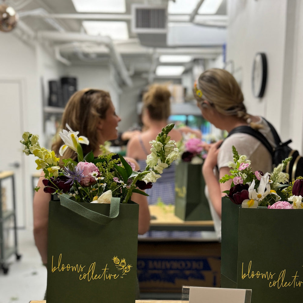 A picture of people standing at a workbench focusing on flower arranging and talking. Two Blooms Collective gift bags in green full of flower bouquet sits at the bottom of picture
