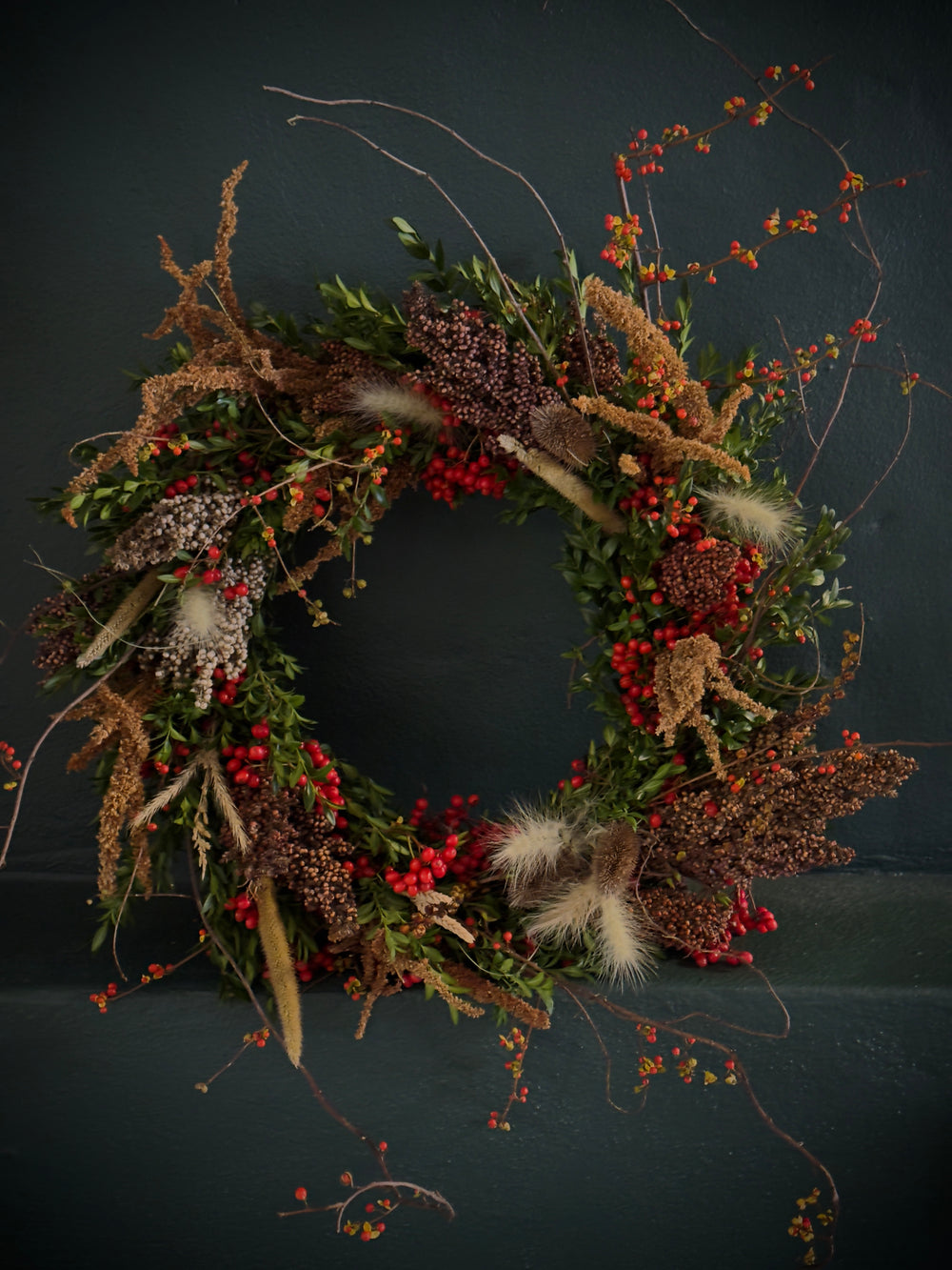 Harvest wreath full of grains, grasses, and berries rests against a dark green wall