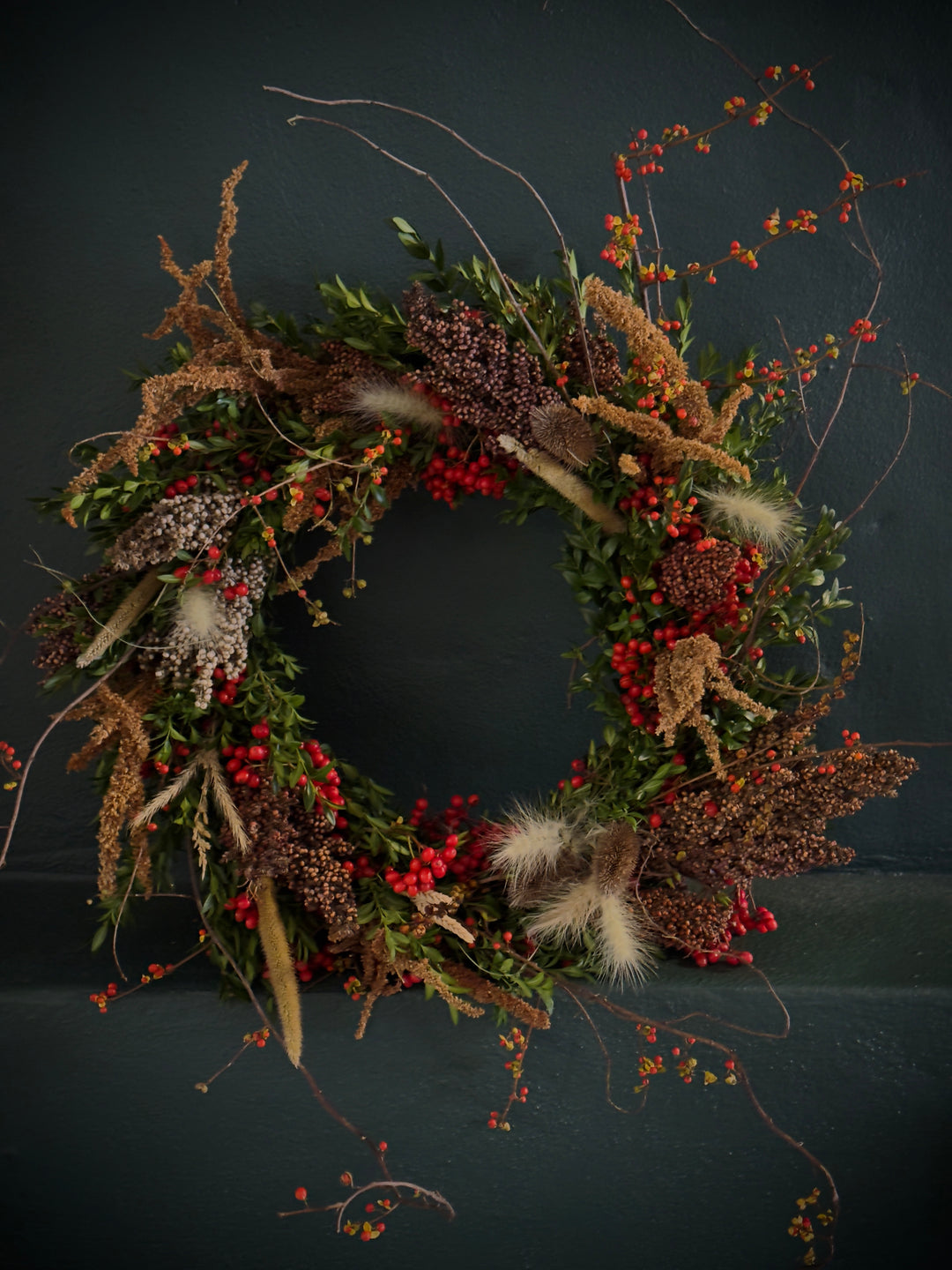 Harvest wreath full of grains, grasses, and berries rests against a dark green wall