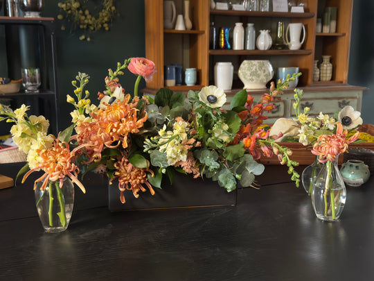 Floral arrangements in vases on a dark surface with a bookshelf in the background