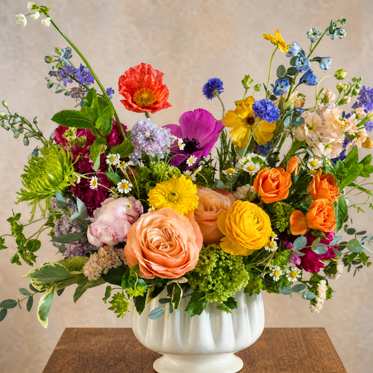 Brightly colored flower arrangement in an off white vase sitting on a wooden table.