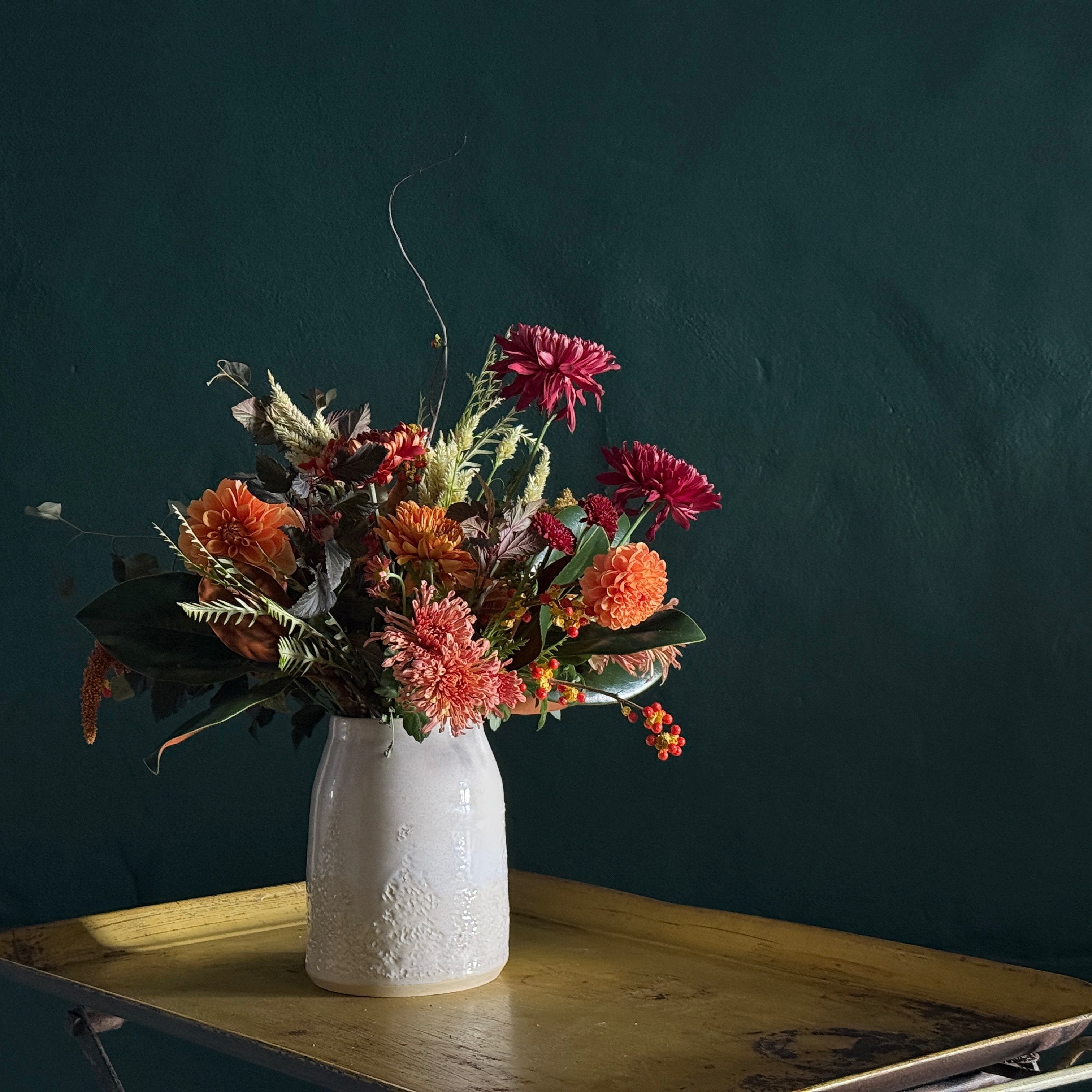 a large flower arrangement of a mix of seasonal blooms in peaches, greens, oranges and burgundies. All arranged in a wide-mouth, off white, ceramic vase