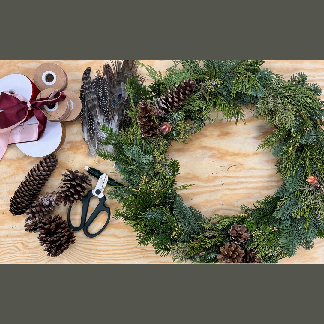 A workshop setup for making holiday wreaths, featuring a 18” grapevine wreath base, greens, wire, ribbons, pinecones, and scissors on a wooden table.
