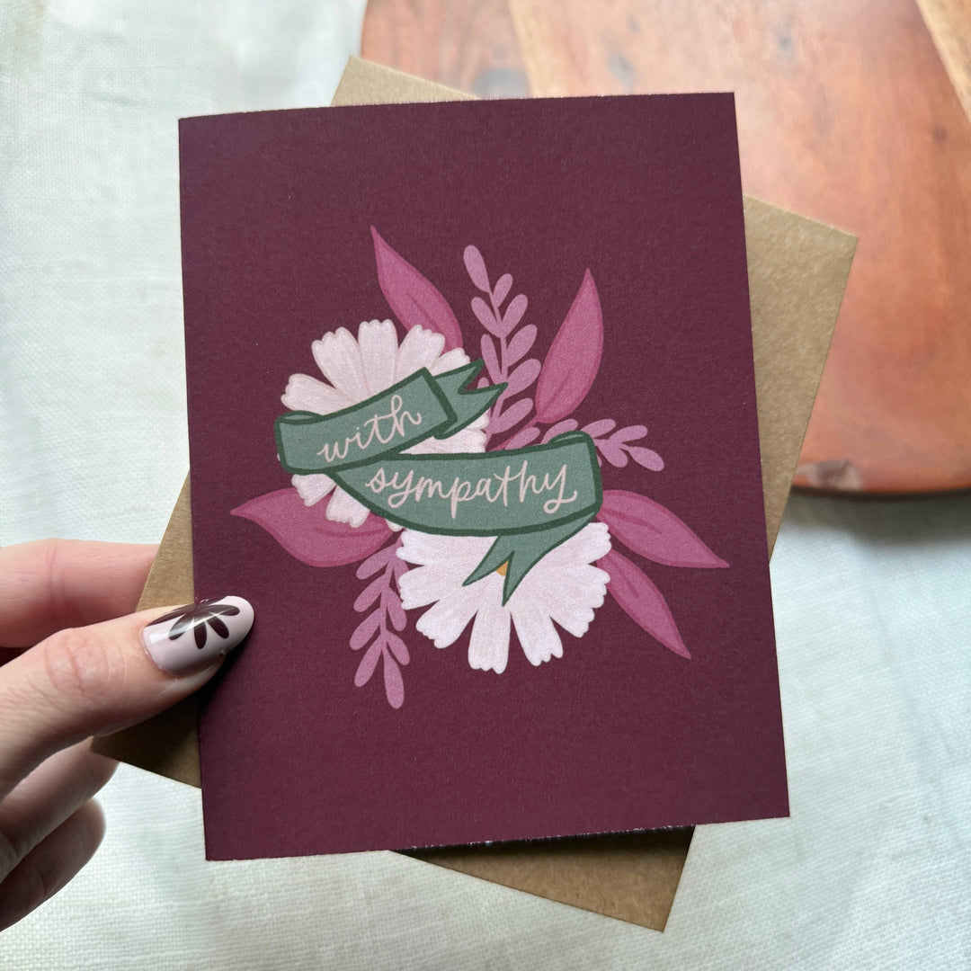 Card with floral design and 'with sympathy' text held by a hand.
