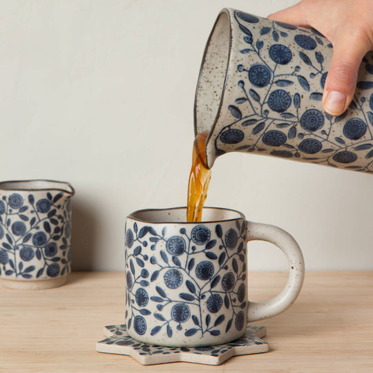 Person pouring coffee from a patterned ceramic mug into another similar mug on a wooden surface.