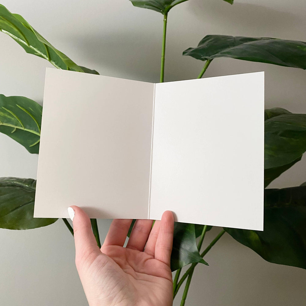 Hand holding a blank white card with green leaves in the background